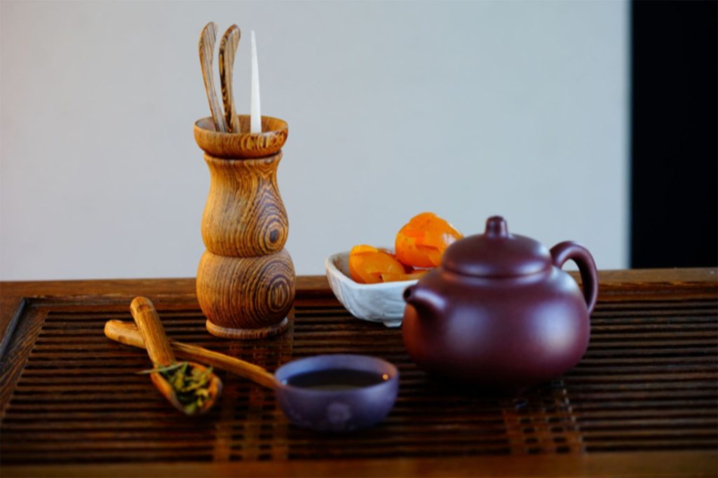 Traditional Chinese tea ceremony set on a slatted wooden tray, featuring a purple clay Yixing teapot, small tea cup, wooden tool holder, bamboo tea scoop with loose leaf tea, and a white dish of dried persimmons