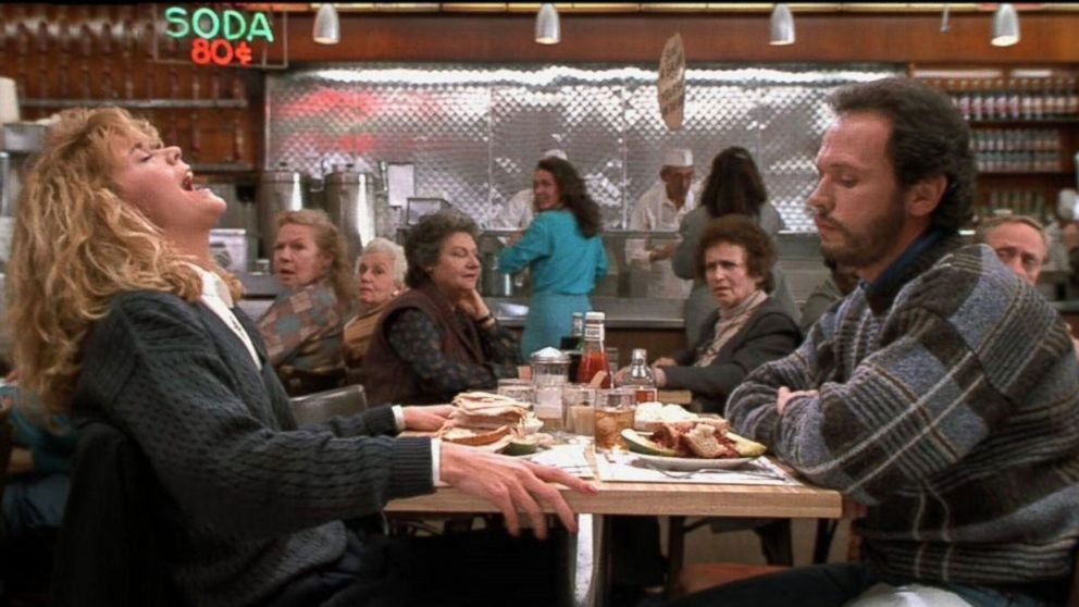 A woman laughing loudly at a diner table while a man sits across from her looking on, surrounded by other restaurant patrons reacting with curiosity