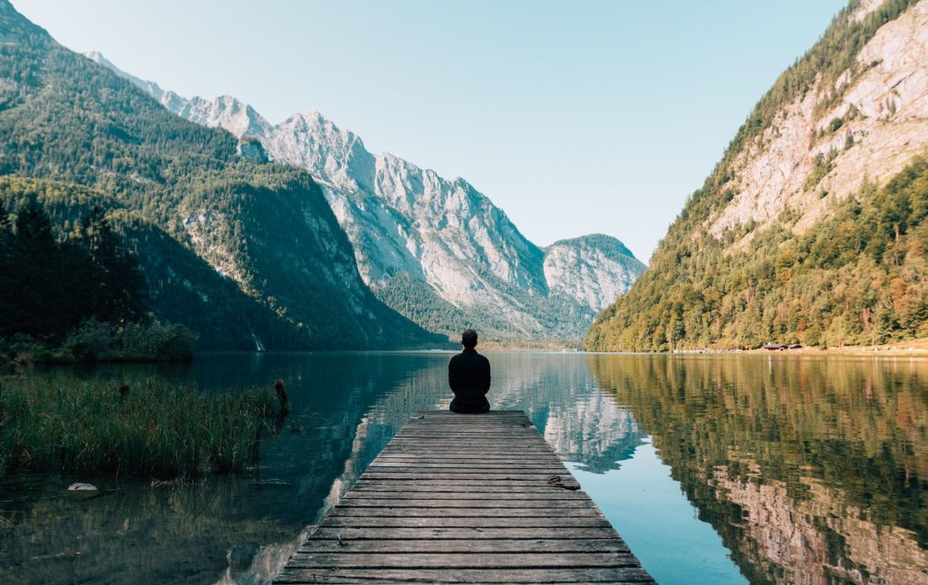 Person seated in meditation at the end of a wooden dock, facing a calm alpine lake surrounded by forested mountains reflected in the still water