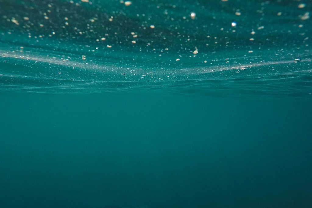 Underwater view looking up through clear teal ocean water toward the shimmering surface, evoking depth, calm, and the subconscious mind