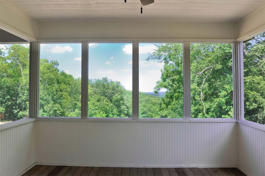 An empty white-paneled screened porch with large windows overlooking a lush green tree canopy under a bright summer sky