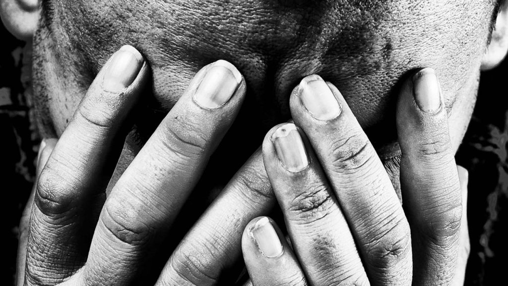 Black and white close-up of a man covering his face with both hands, conveying grief or emotional overwhelm