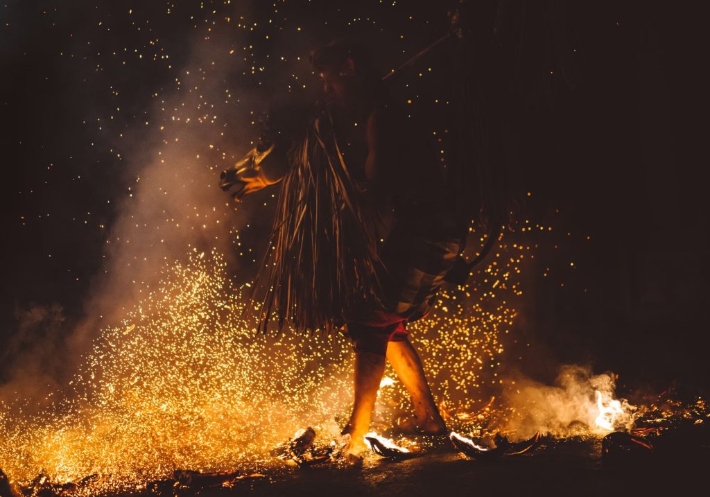 Figure in a traditional grass skirt dancing amid a shower of fire sparks and smoke in a nighttime ritual ceremony